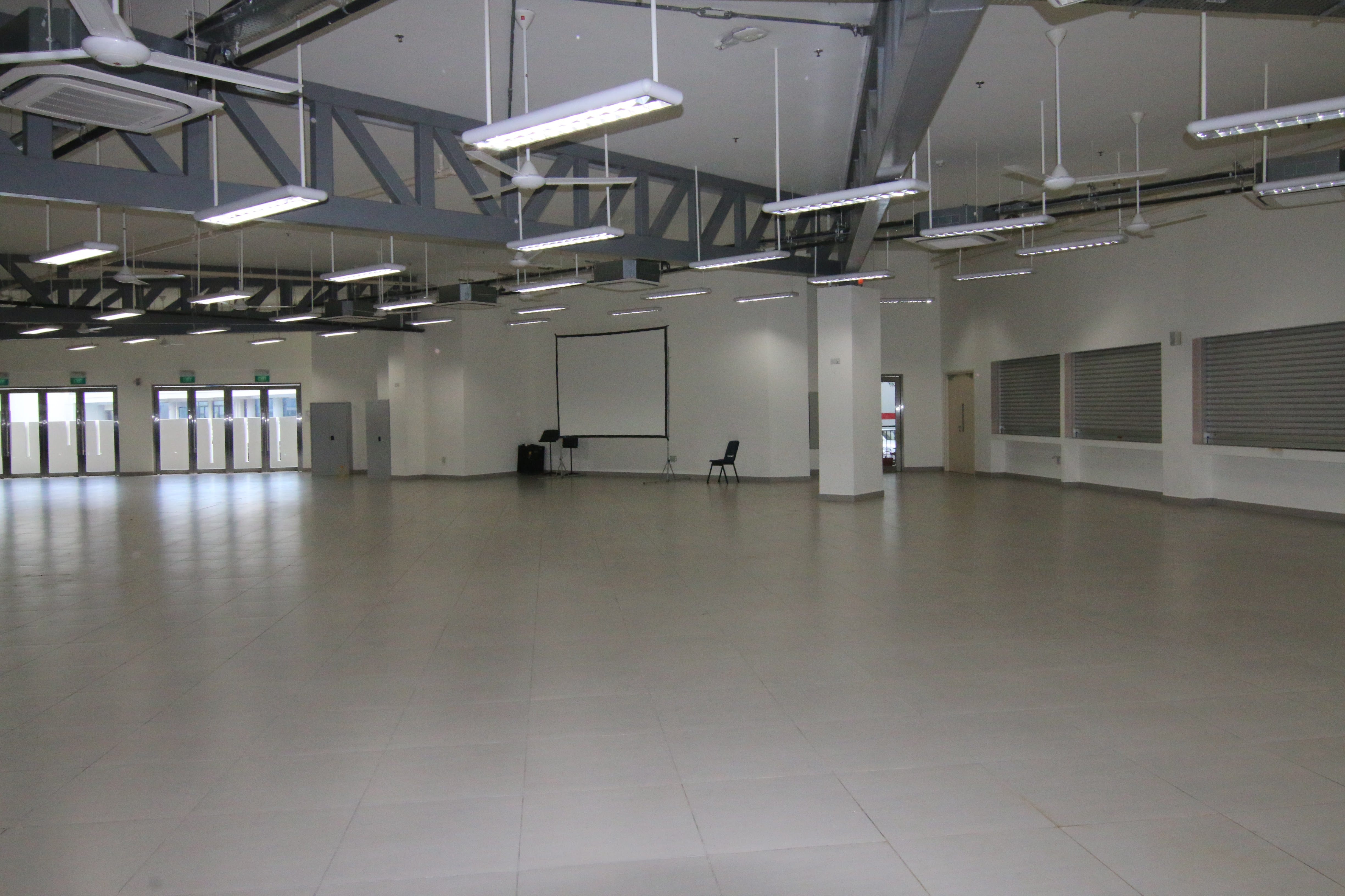 Large empty industrial room with exposed ceiling trusses, fluorescent lights, a projector screen, and a smooth gray floor.